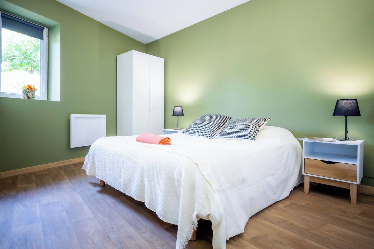 A bedroom with a bed, nightstands, a lamp, and a window with a view of greenery.