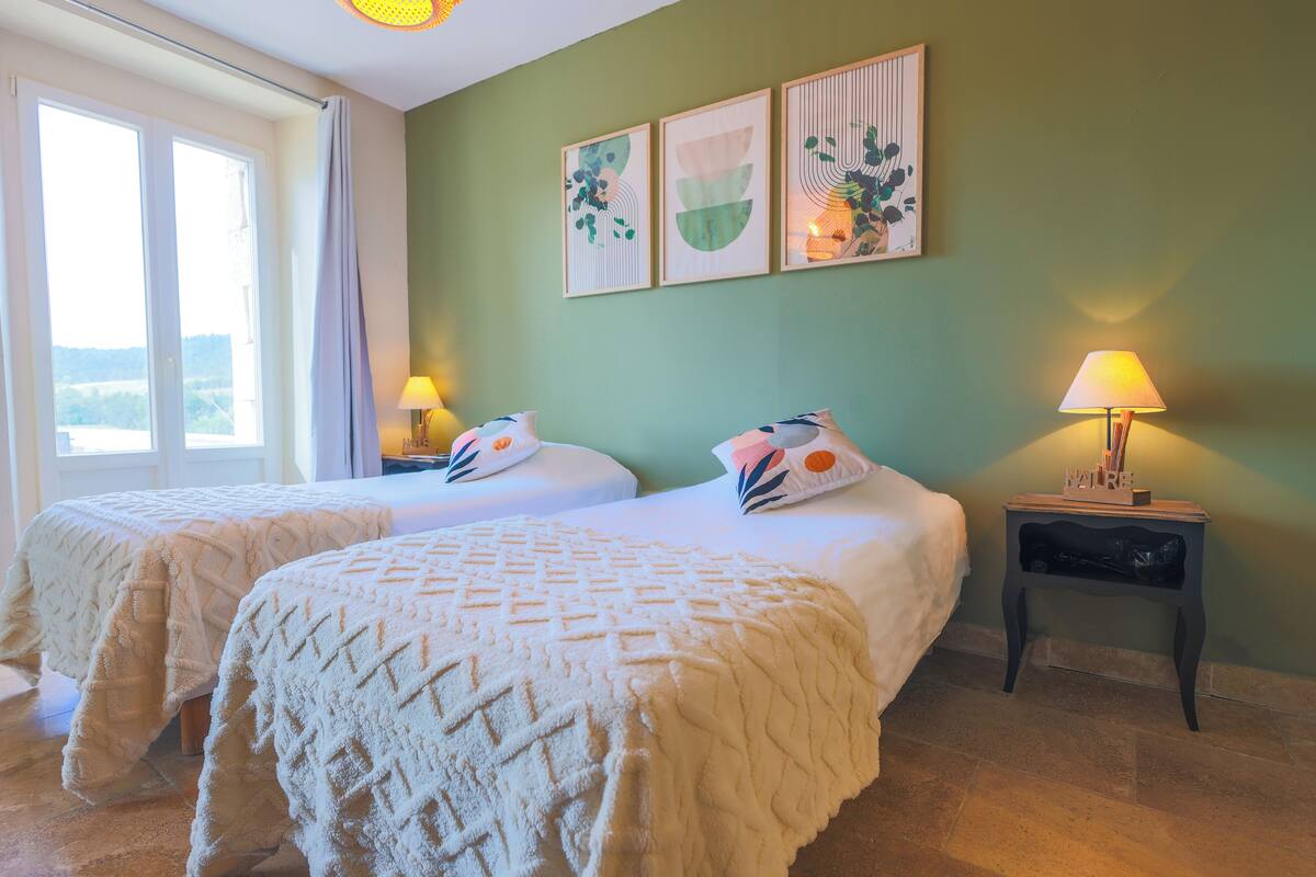 A room with two beds, a nightstand with a lamp, and a window with curtains.