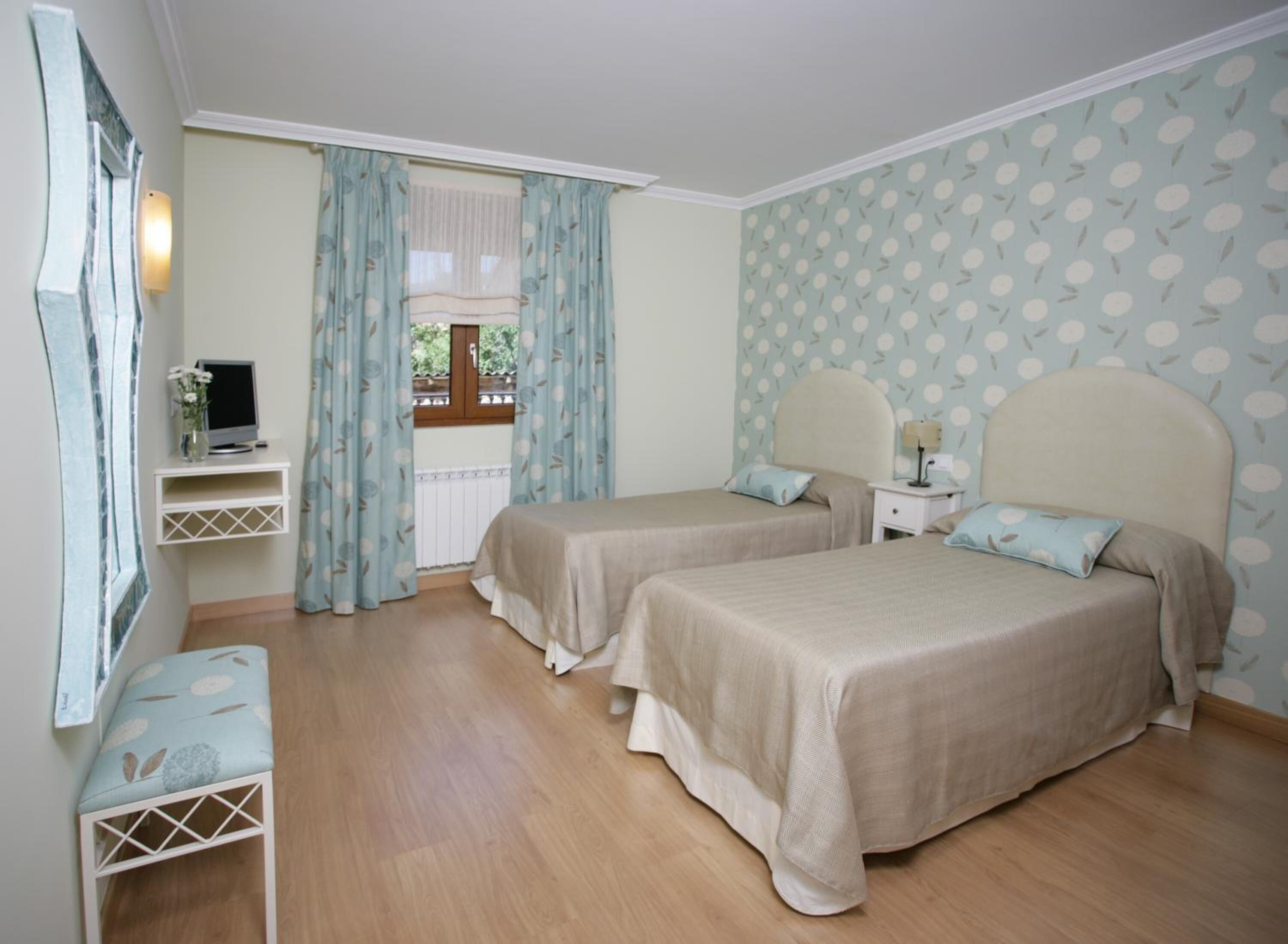 A bedroom with two beds, a TV on a stand, a small table, and a window with curtains.