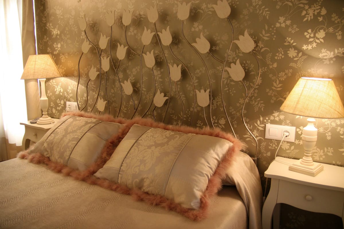 Une chambre à coucher avec un papier peint à motifs floraux, deux lampes de chevet et un lit agrémenté d’oreillers à motifs.