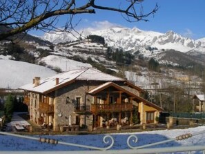 A wooden chalet with a balcony, set against a backdrop of snow-covered mountains. - Posada El Corcal De Liébana (Cillorigo de Liebana)
