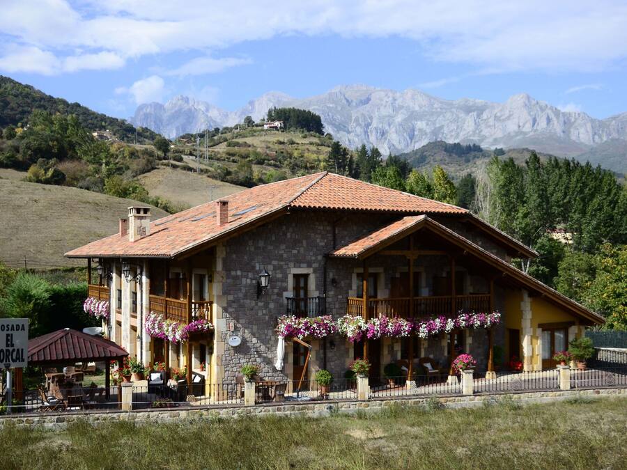 Un edificio de piedra con techo de tejas, un balcón adornado con cestas de flores colgantes y un letrero que dice "POSADA EL CORCAL".