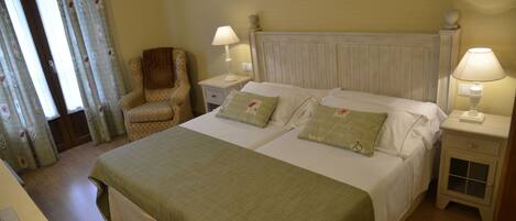 Una cama doble con una colcha verde, dos mesitas de noche con lámparas, una silla y una ventana con cortinas.