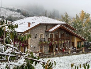A stone building with a sloped roof, a balcony with hanging plants, and a snow-covered ground. - Posada El Corcal De Liébana (Cillorigo de Liebana)