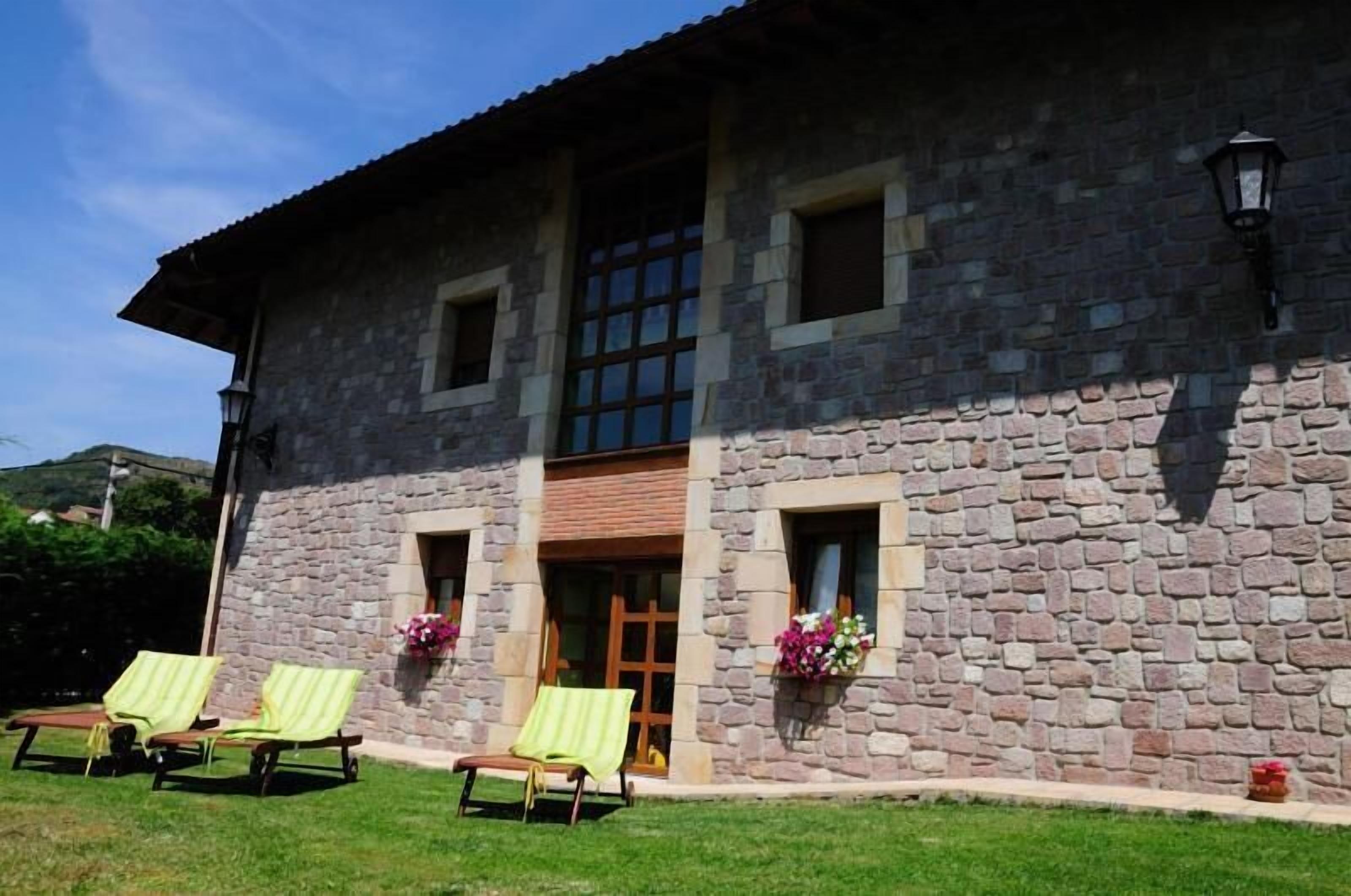 A stone building with a tiled roof, a wooden door, and two windows. There are two lounge chairs with yellow cushions outside.