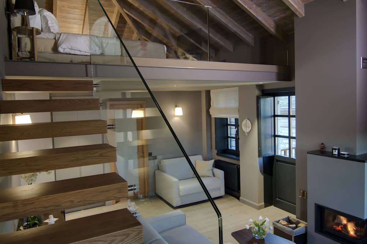 A spacious loft with a high ceiling, exposed wooden beams, a staircase, a living area with a sofa, a fireplace, and a dining table.