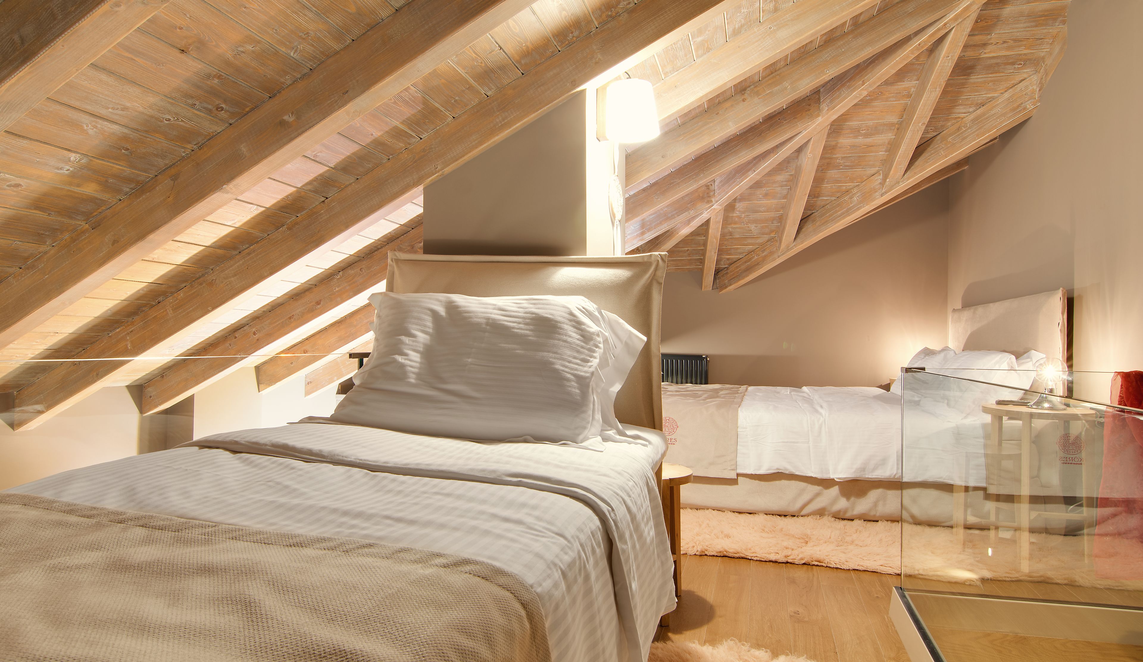 Un dormitorio con techo inclinado, una cama con ropa de cama blanca, una mesita de noche y una lámpara.
