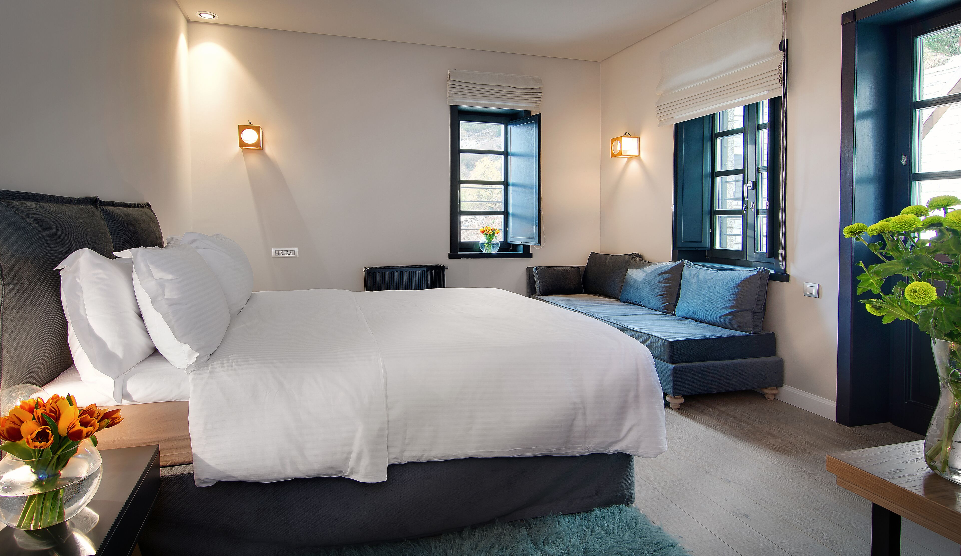A modern bedroom with a large bed, a bedside table with a vase of flowers, a window with a view, and a blue door.