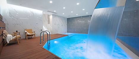 A modern indoor pool area with a wooden deck, lounge chairs, and a water feature.