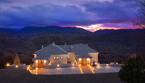 A stone house with a stone driveway, surrounded by mountains and a sunset sky.