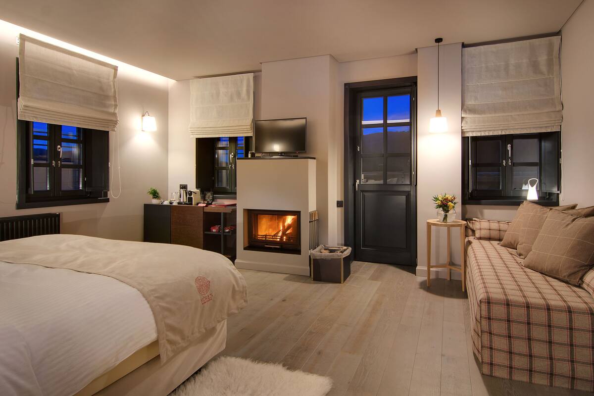 A bedroom with a bed, a sofa, a fireplace, a TV, and a door with a view of the outside.
