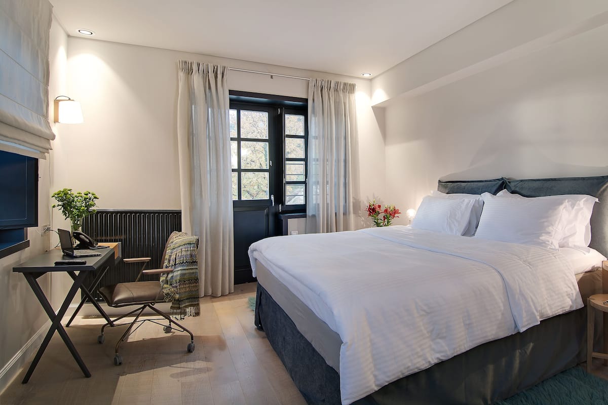 A modern bedroom with a large bed, a desk with a laptop, a chair, and a small table with a lamp.