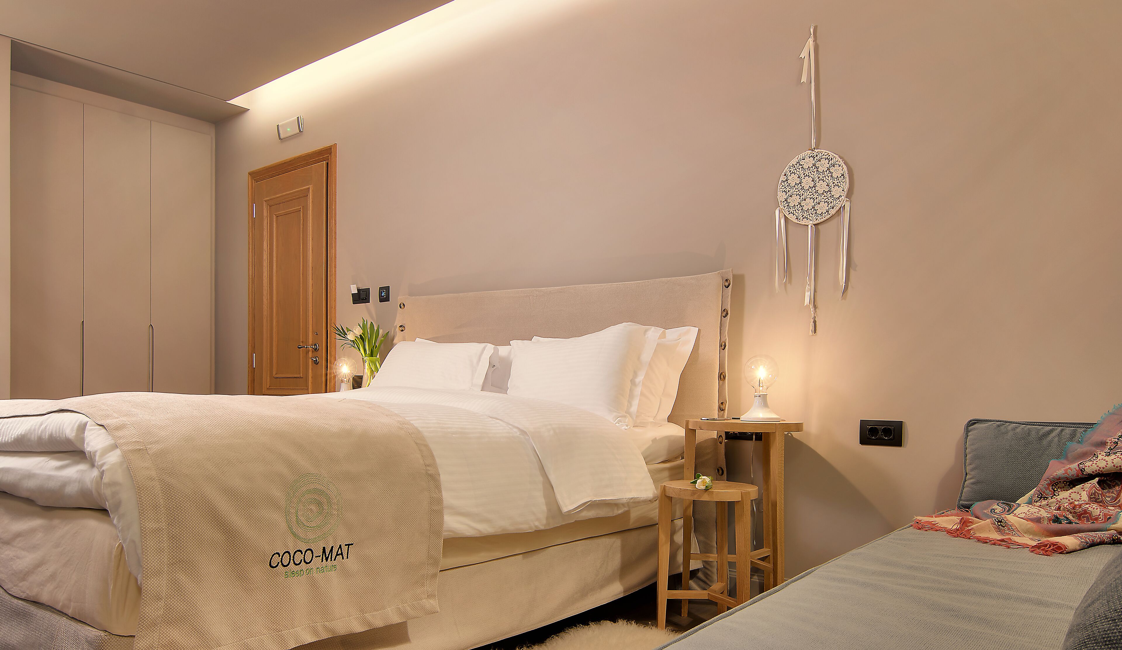 Een slaapkamer met een bed, nachtkastje, lamp en een decoratief hangobject.