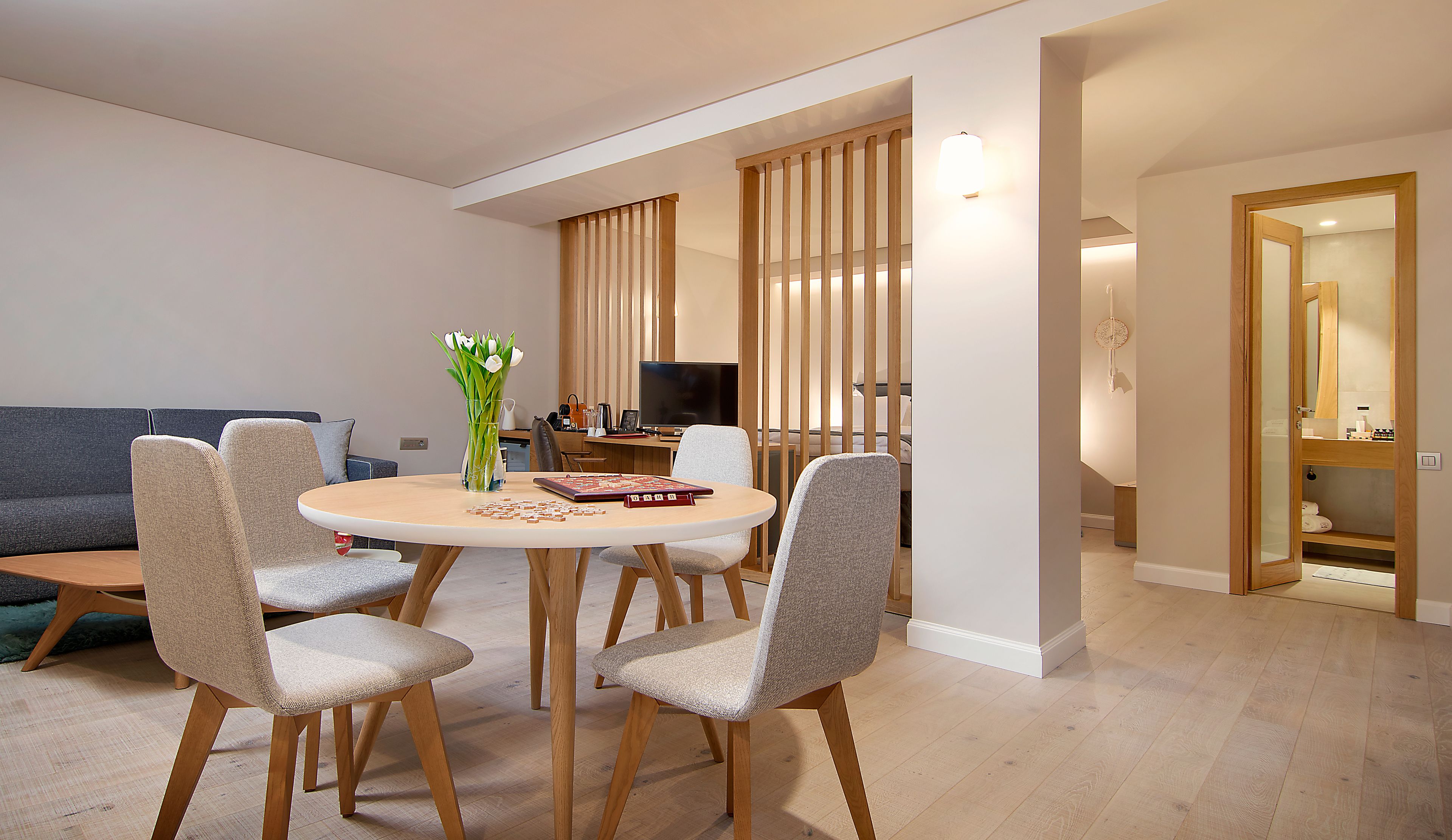 A modern living room with a round dining table, four upholstered chairs, a grey sofa, and a wooden TV stand.