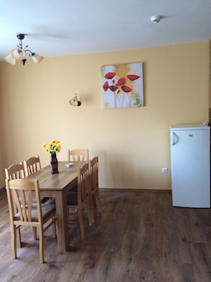 A dining area with a wooden table and chairs, a vase with flowers, a ceiling fan, a wall-mounted air conditioning unit, and a refrigerator. - Horizont Apartments (Sunny Beach)