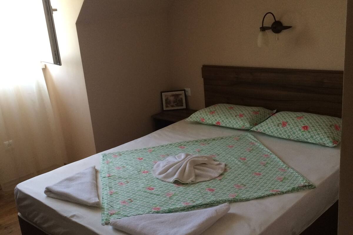 Un lit bien fait, recouvert d’une couvre-lit à motifs floraux et agrémenté de deux oreillers, une table de chevet avec une lampe et un cadre photo accroché au mur.