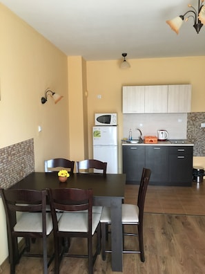 A compact kitchen with a microwave, refrigerator, and sink. There is a dining table with chairs and a vase with flowers. - Horizont Apartments (Sunny Beach)