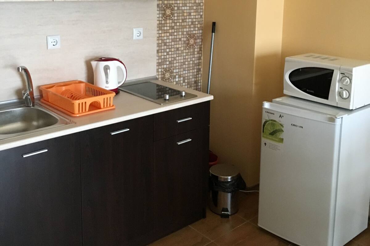 Une cuisine compacte avec des meubles de couleur sombre, un plan de travail blanc, un four à micro-ondes, une bouilloire et un évier.