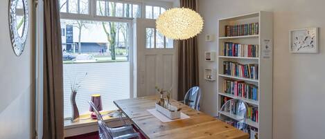 A dining area with a wooden table, clear chairs, a hanging light fixture, a bookshelf, and a window with curtains.