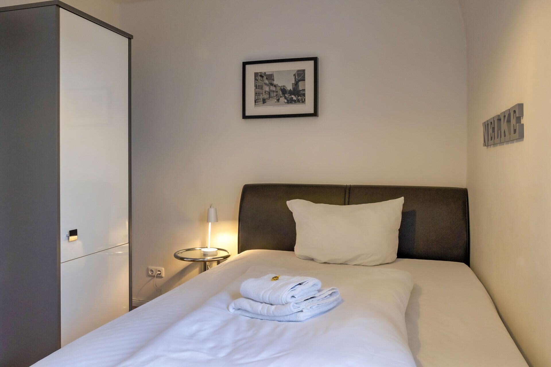 A hotel room with a bed, a headboard, a bedside table with a lamp, a framed picture on the wall, and a wardrobe.