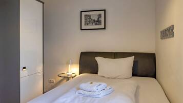 A hotel room with a bed, a headboard, a bedside table with a lamp, a framed picture on the wall, and a wardrobe.