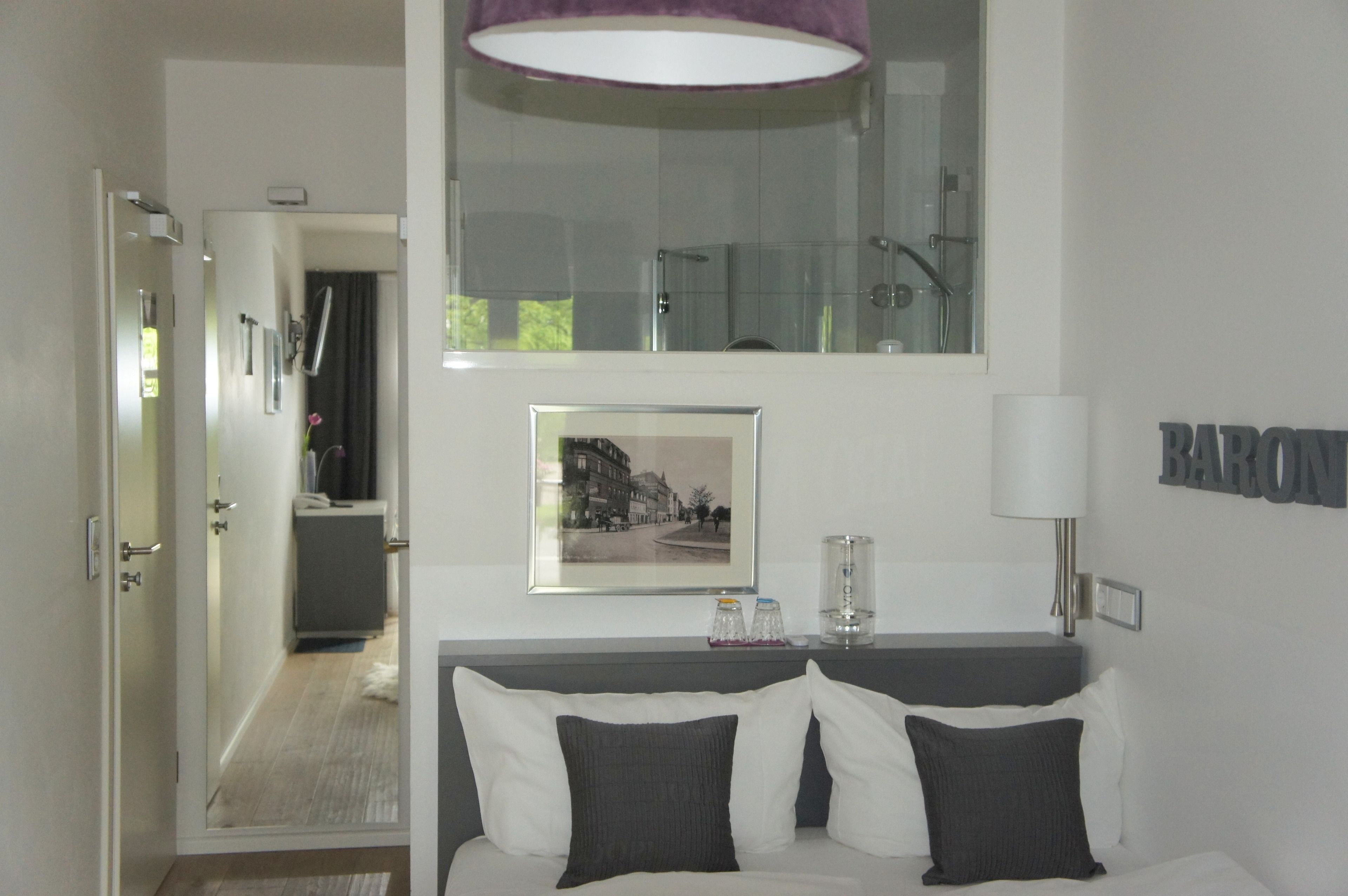 Una habitación de hotel con una cama, una mesita de noche, un espejo, un cuadro enmarcado, una lámpara y un baño visible a través de una puerta abierta.