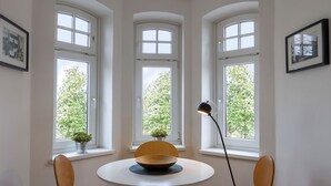 A dining area with a round table, four chairs, and a floor lamp. Three windows provide natural light and a view of greenery outside.