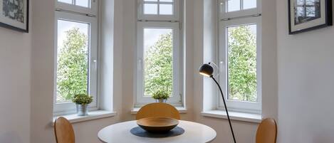 A dining area with a round table, four chairs, and a floor lamp. Three windows provide natural light and a view of greenery outside.