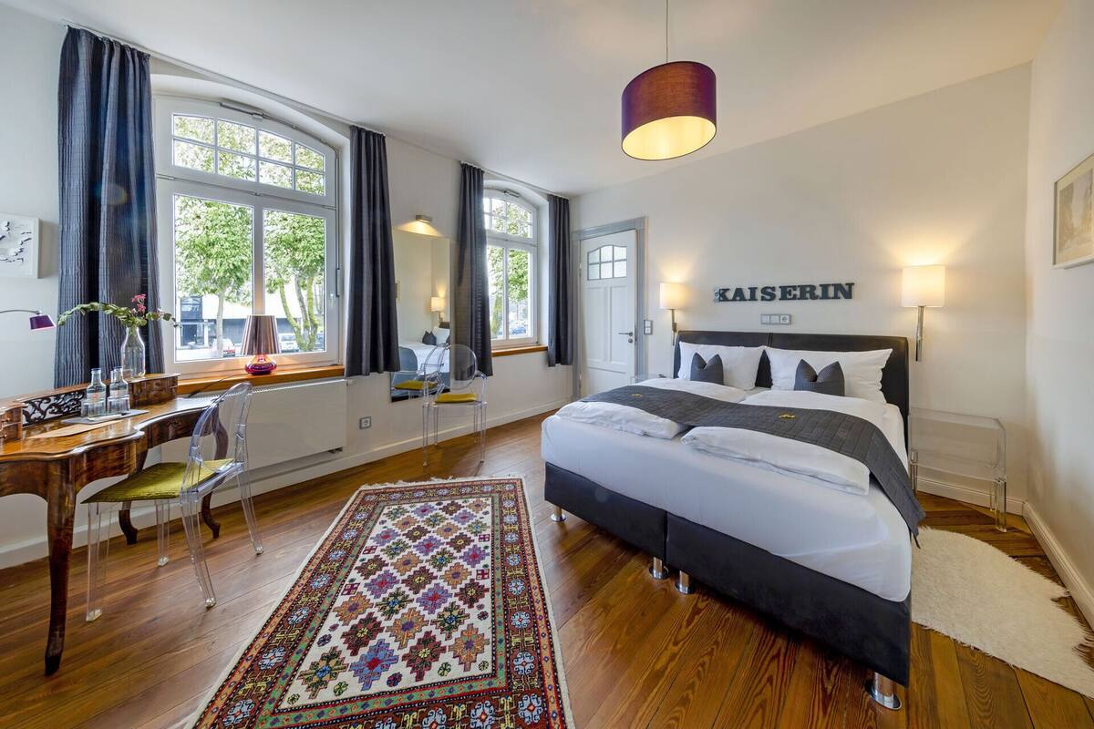 A hotel room with a large bed, a desk with a chair, a window with curtains, and a rug.