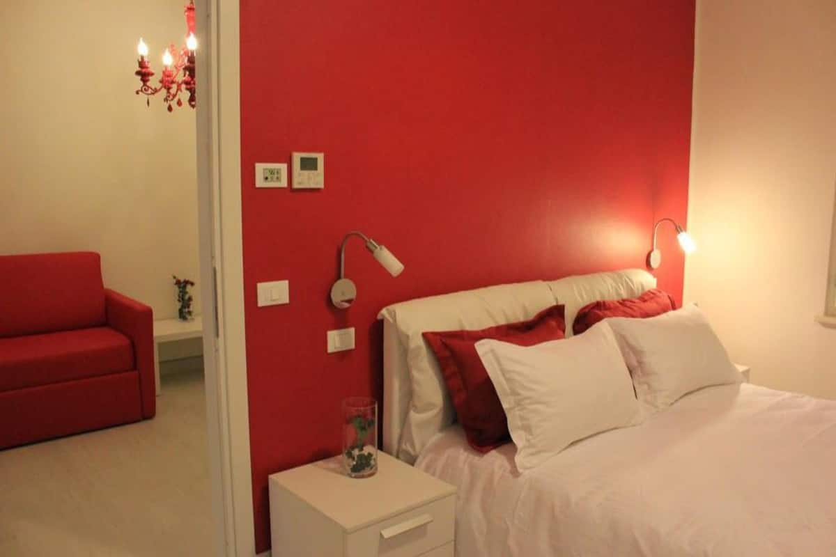 Une chambre avec un mur d’accent rouge, un lit blanc, un canapé rouge, une table de chevet avec une lampe et une commode avec une lampe.