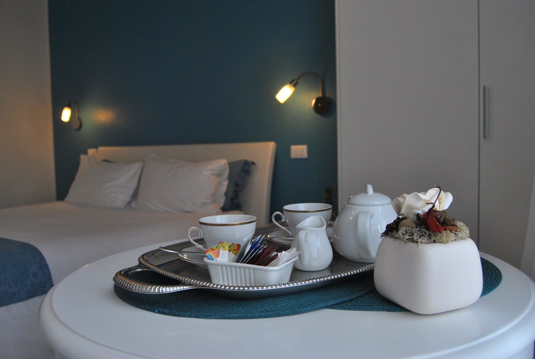 A neatly arranged breakfast tray with cups, a pitcher, and a decorative vase on a round table in a hotel room with a bed and wall-mounted lights.