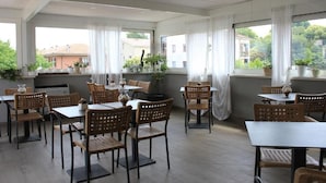 Un vaste espace dédié à la salle à manger, avec des chaises et des tables en bois, de grandes fenêtres ornées de rideaux blancs et des plantes en pot.