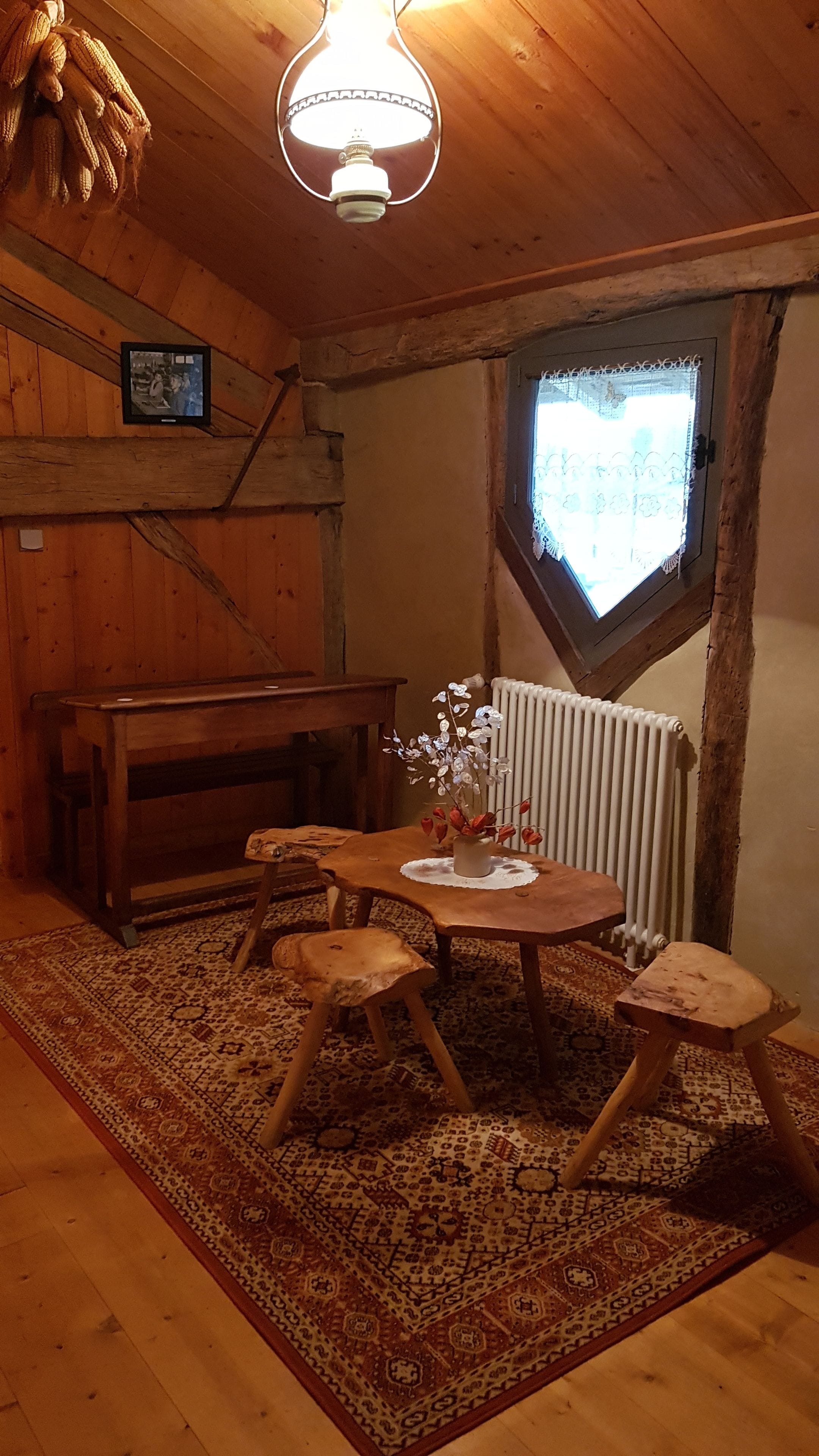 Una habitación rústica con techo de madera, una mesa de madera con cuatro taburetes, un radiador y una ventana con cortinas de encaje.