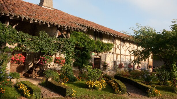 Un edifici tradicional amb teulades ceràmiques, una torre campanar i un jardí ple de flors i arbustos diversos.