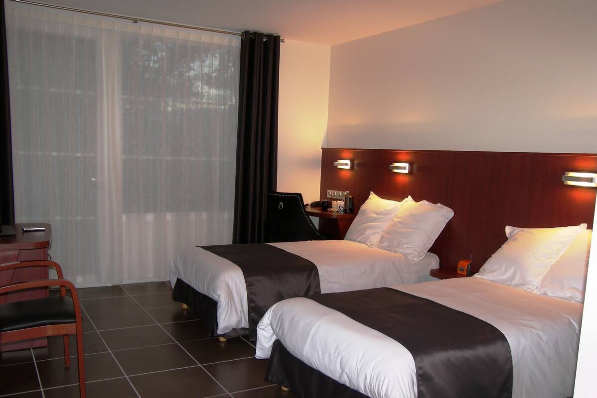 Quarto de hotel com duas camas, uma escrivaninha com cadeira e uma janela grande com cortinas.