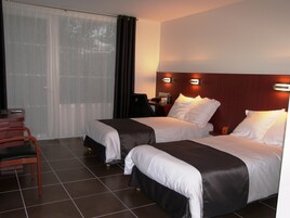 Quarto de hotel com duas camas, uma escrivaninha com cadeira e uma janela grande com cortinas.