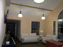 Uma sala de estar com dois sofás brancos, teto de madeira, uma janela grande com persianas e uma televisão fixada na parede.