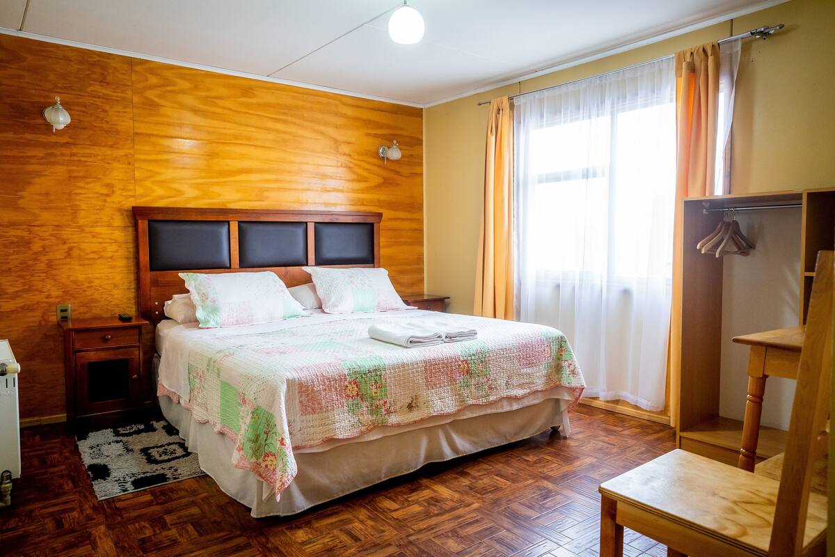 A bedroom with a wooden headboard, a bed with a floral bedspread, a bedside table, a nightstand, a wooden chair, and a wardrobe.