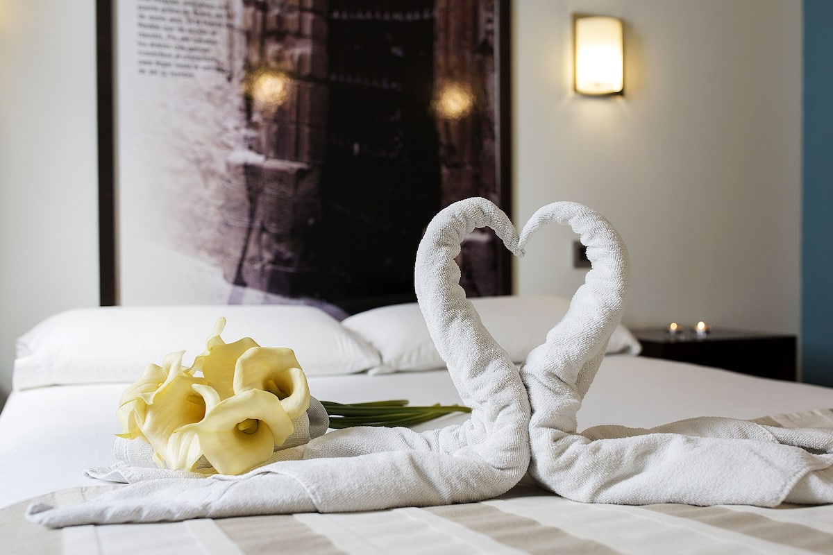 Una cama bien hecha con una toalla dispuesta en forma de corazón y un ramo de flores amarillas.