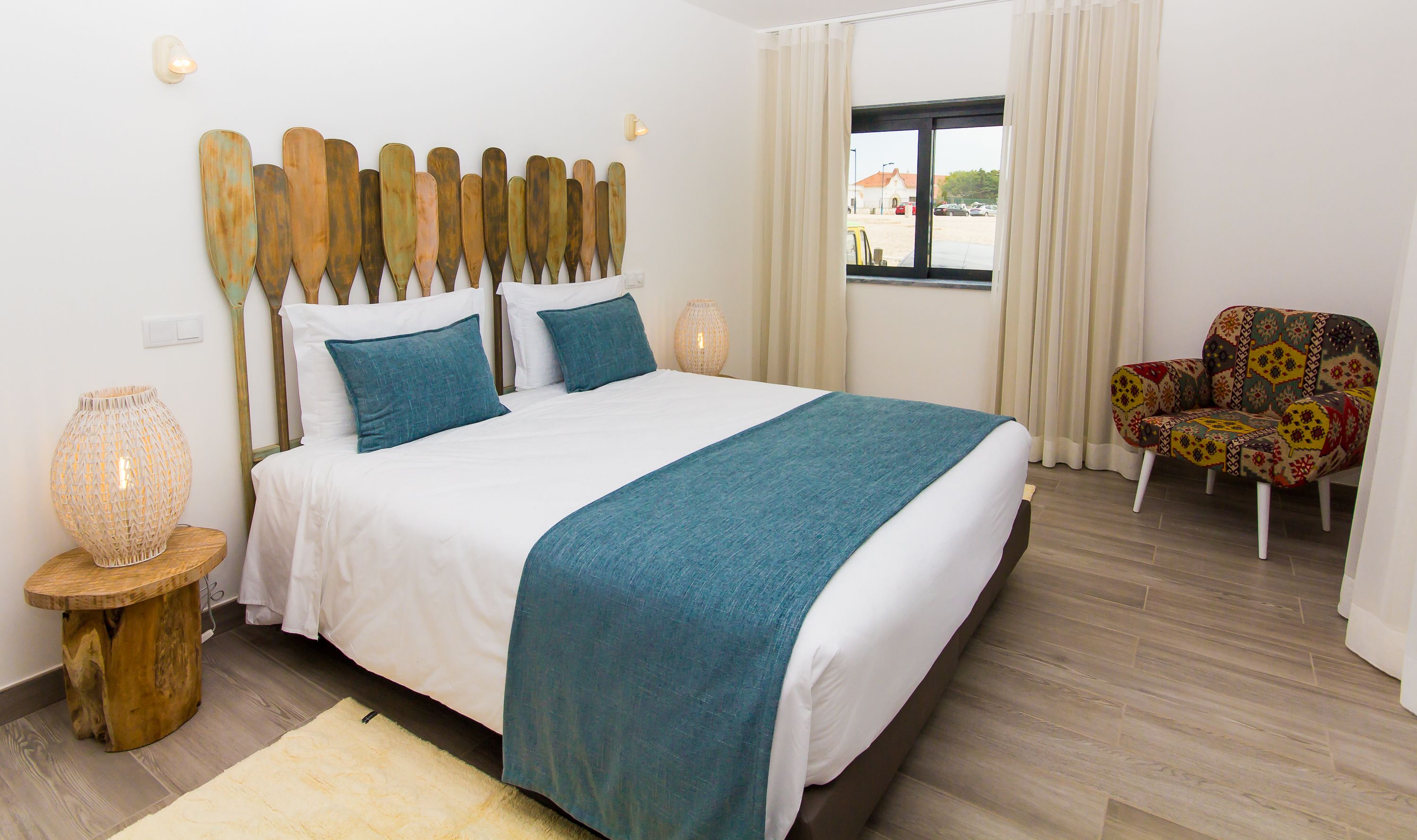 Un dormitorio ordenado con cama, dos almohadas, lámpara de noche, una silla y ventana con cortinas.