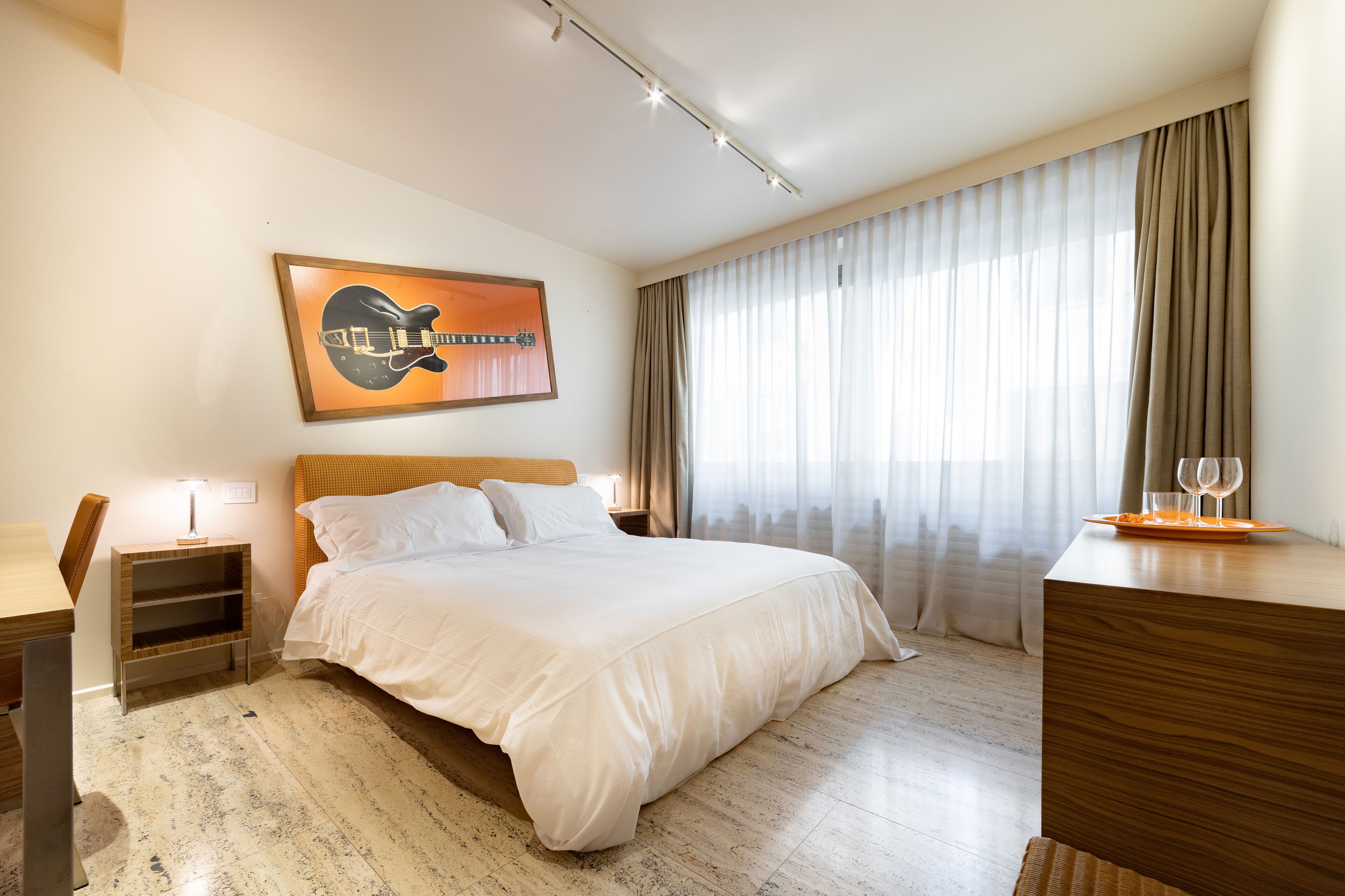 A hotel room with a bed, a desk, a chair, a nightstand, a lamp, a painting, and a window with curtains.