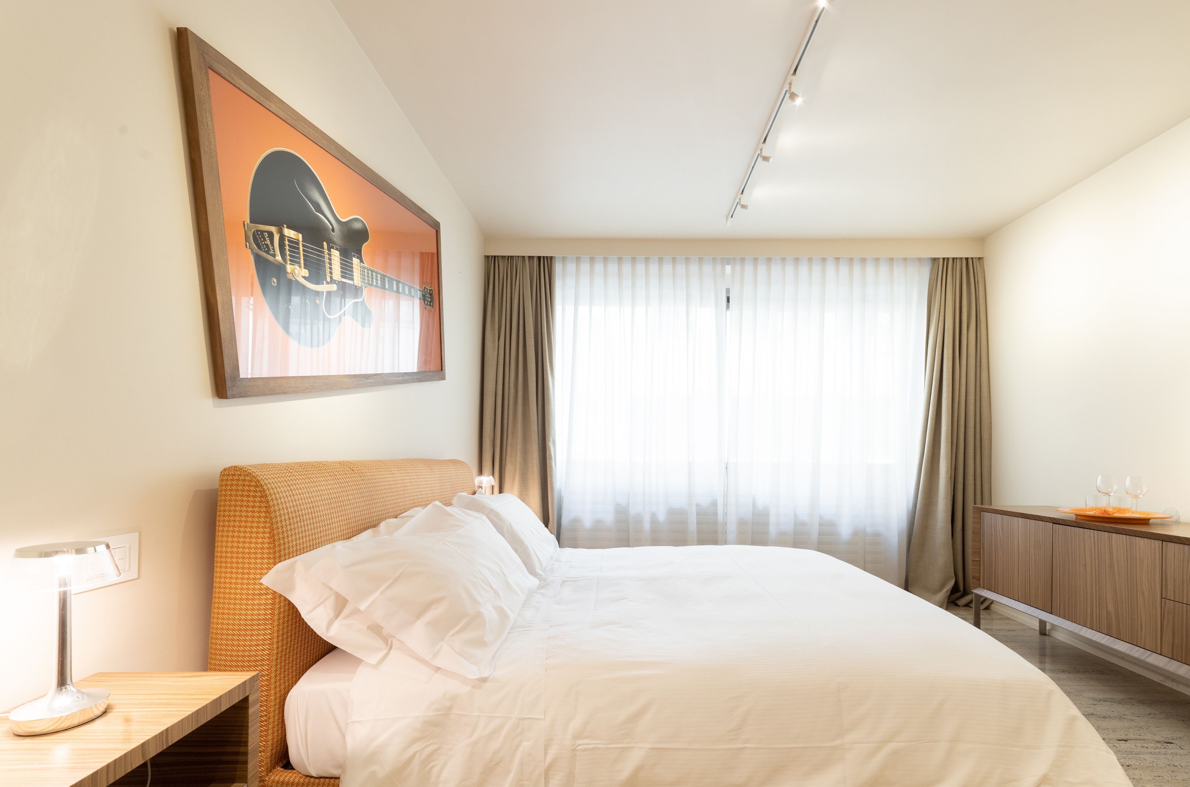 Ein Hotelzimmer mit einem großen Bett, einem Nachttisch mit Lampe, einem Holz-Kopfteil, einem gerahmten Bild an der Wand, einem Fenster mit Vorhängen und einer Holzkommode mit einem Tablett mit Gläsern.