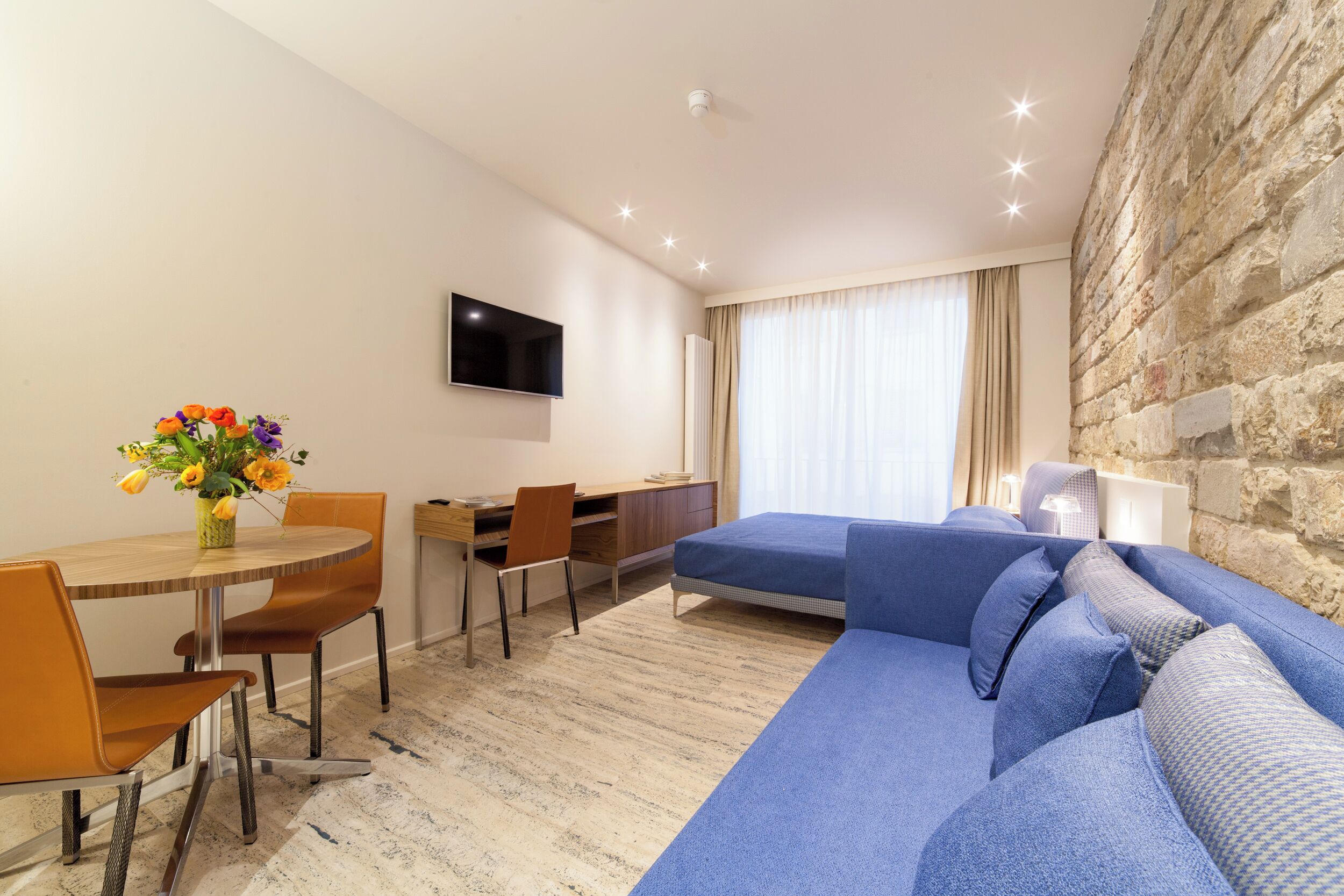 A modern hotel room with a dining area, a flat-screen TV, a sofa, and a stone accent wall.