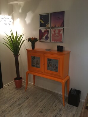 A room with a wooden floor, a potted plant, an orange cabinet with artwork, and a wall-mounted light fixture. - Le stanze degli amidei (Florence)