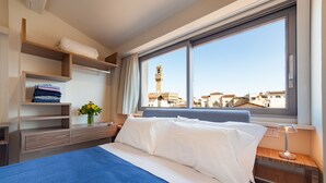 A hotel room with a bed, a nightstand, a window with a view of a tower, and built-in shelves with folded towels. - Le stanze degli amidei (Florence)