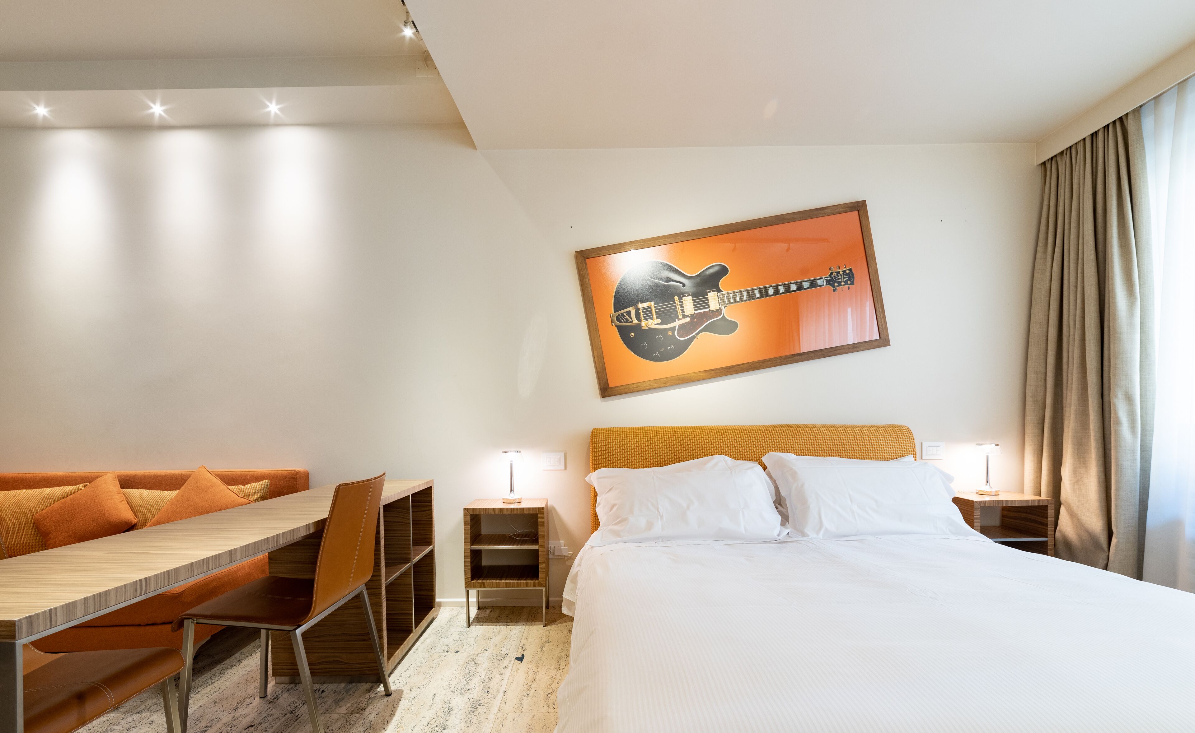 Ein Hotelzimmer mit Bett, Schreibtisch, Stuhl, einem Gitarrenbild an der Wand und einem Fenster mit Vorhängen.