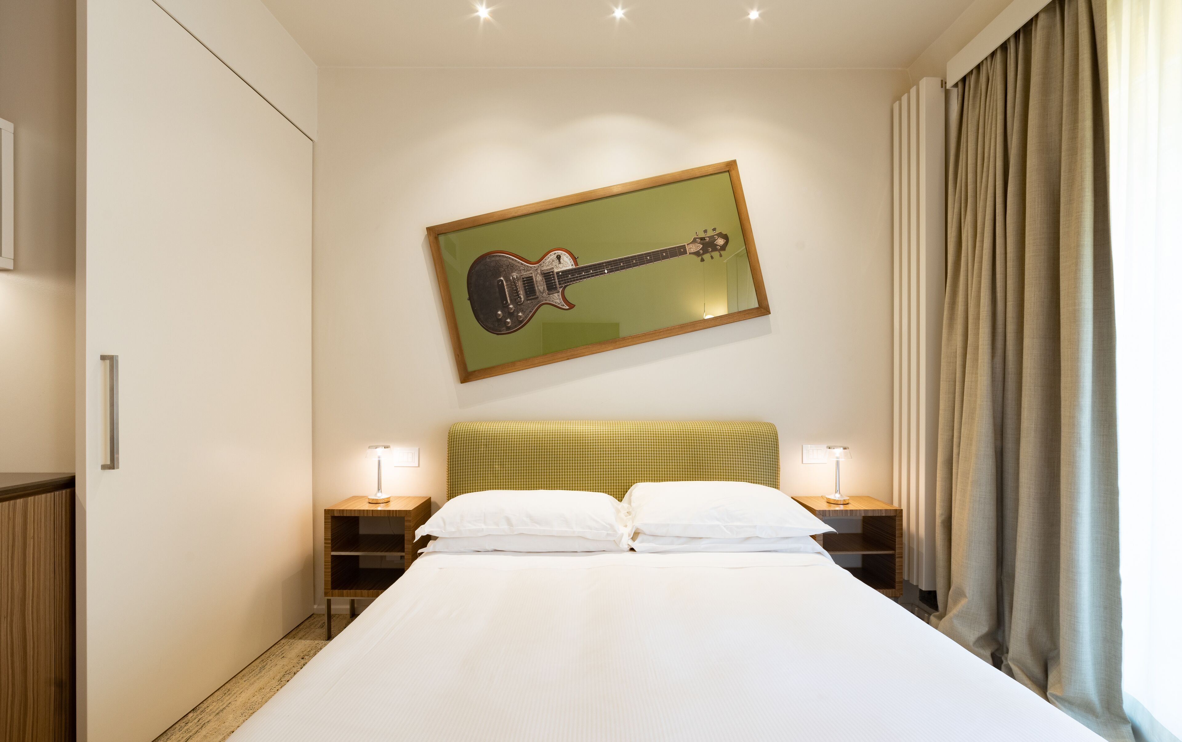 A hotel room with a bed, two bedside tables, a guitar artwork on the wall, and a window with curtains.