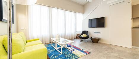 A modern living room with a bright yellow sofa, a coffee table, a colorful rug, a flat-screen TV mounted on the wall, and a large window with sheer curtains.
