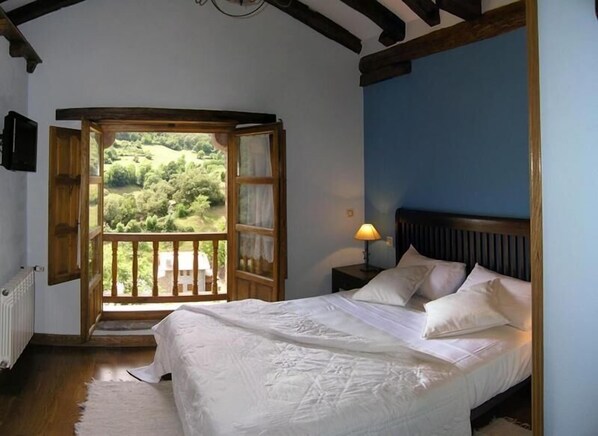 A bedroom with a bed, a nightstand, a lamp, and a view of a green landscape through an open window.