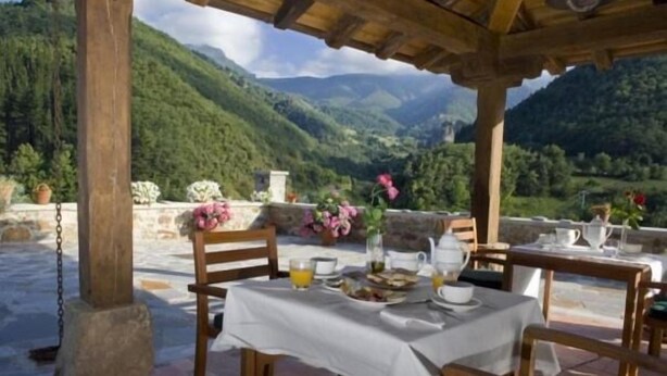 An outdoor dining area with a table set for two, surrounded by wooden chairs, overlooking a mountainous landscape.